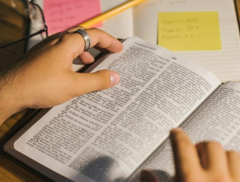 Close-up of hands holding and reading a Bible with study notes and a pencil on a desk.